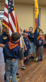Seven Cub Scouts in uniforms are holding up an american and pack flags on poles. They are getting ready to perform a flag ceremony.