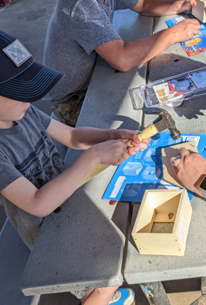 A Tiger Cub Scout holding a hammer while an adult holds the nail. They are building a bird house on a picnic table with blue instructions underneath the birdhouse
